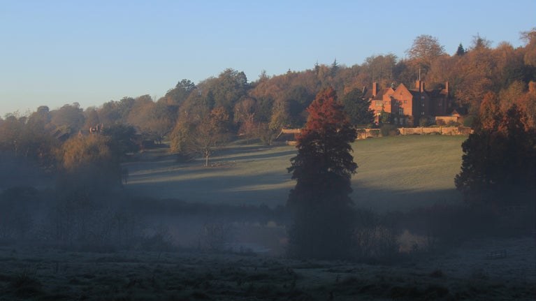 A view from across the lake to the house at Chartwell, Kent, at the top of a slope dotted with trees with mist rising off the lake